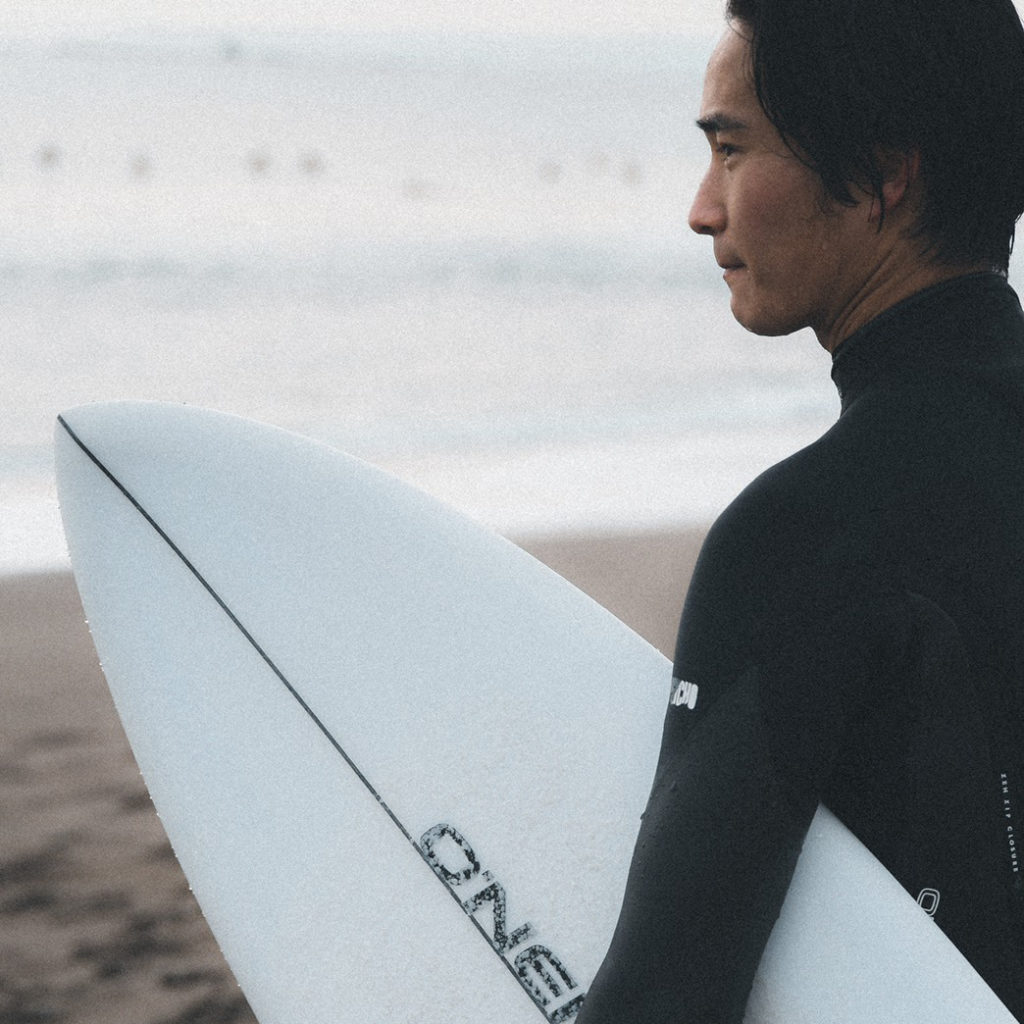 O'NEILL（オニール）チームメンバー AKITO MATSUNO（まつの あきと） サーフボード片手に砂浜に立つ横顔